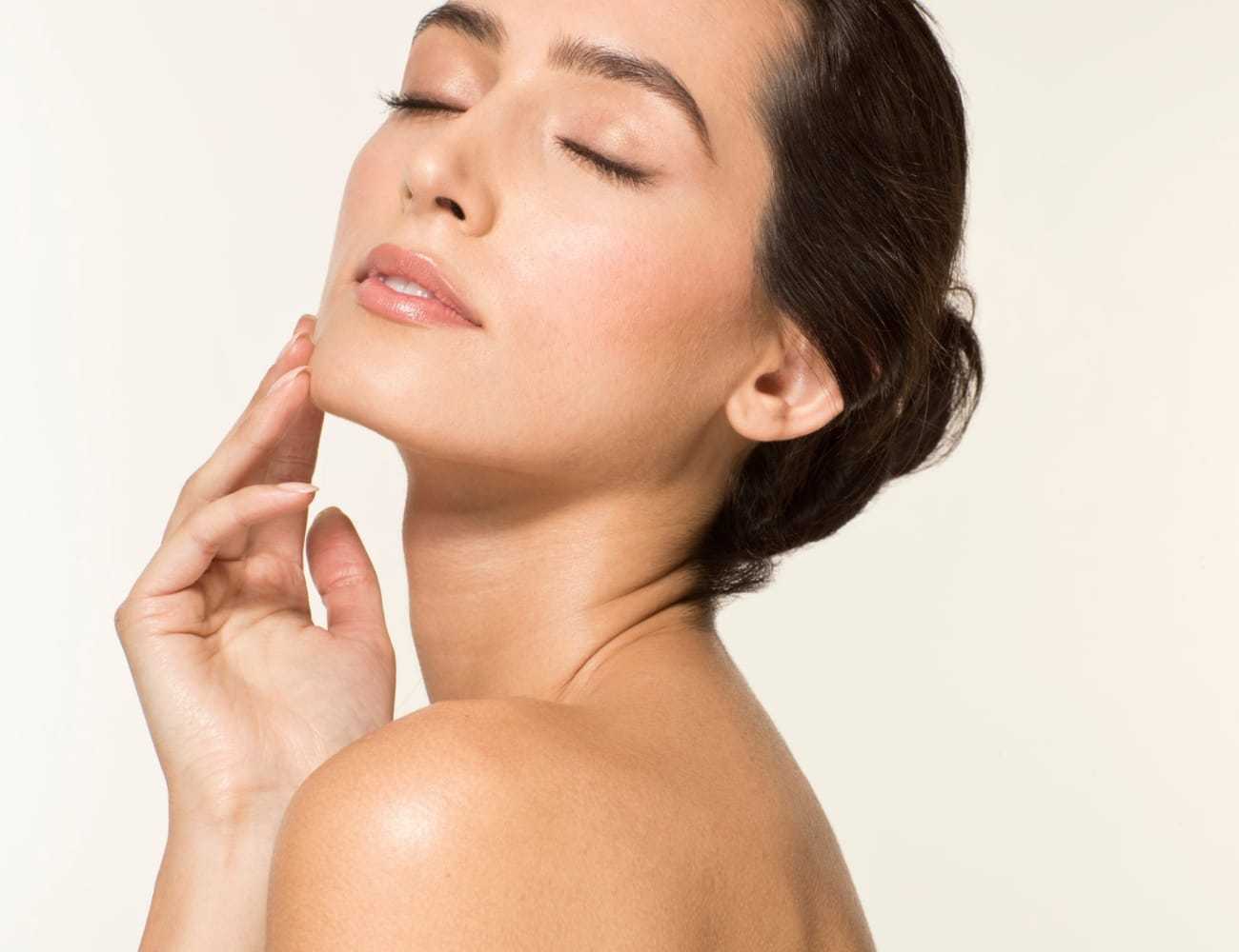Woman with closed eyes, touching her face gently against a soft background.